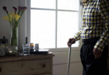 Elderly man standing with a cane next to a table with flowers and medication