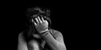 A person sitting in a curled position, covering their face with one hand in a dark setting