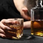 Alcohol’s Hidden Assault on Your Skin A person with their head down, holding a glass of whiskey next to a bottle