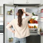 Refrigerator Staples Slash Cholera Risk 100x A woman standing in front of an open refrigerator filled with various fruits and vegetables