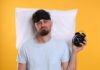 Sleep Patterns Tied to Cancer Risk Man wearing a sleep mask holding an alarm clock with a frustrated expression