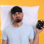 Sleep Patterns Tied to Cancer Risk Man wearing a sleep mask holding an alarm clock with a frustrated expression