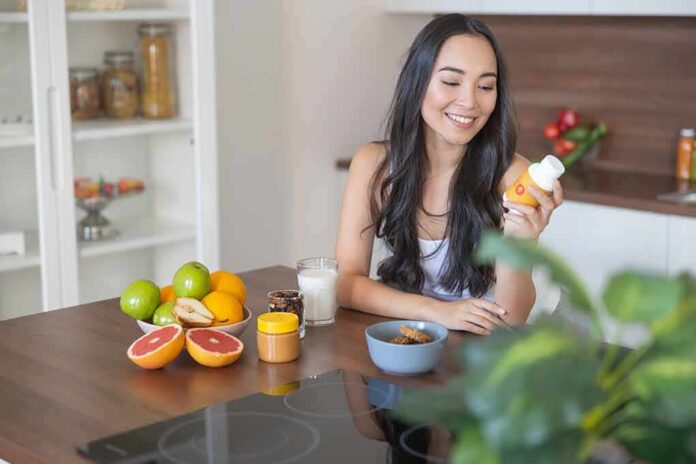 shutterstock_1572303289 Woman sitting at kitchen counter holding a supplement bottle
