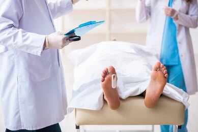 Medical staff stand near a covered body on a gurney with a toe tag