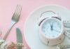 Pink alarm clock on a white plate with fork, knife, and measuring tape