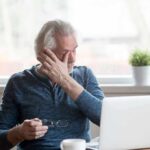 Older man rubbing his eye while sitting at a laptop