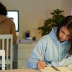 Student in blue hoodie writing in notebook at desk
