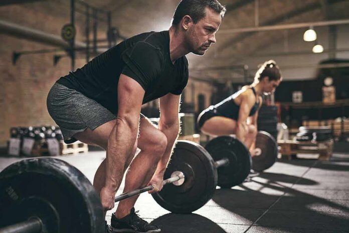 shutterstock_791792701 Man deadlifting a barbell in a gym with a woman lifting behind him