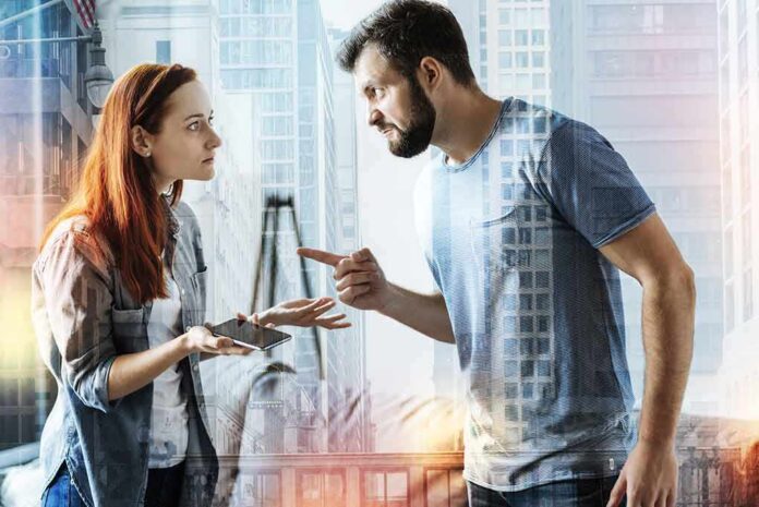shutterstock_1007784238 Two adults arguing indoors, man pointing at woman holding a phone