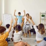 Children raise hands in a classroom circle with teacher clapping
